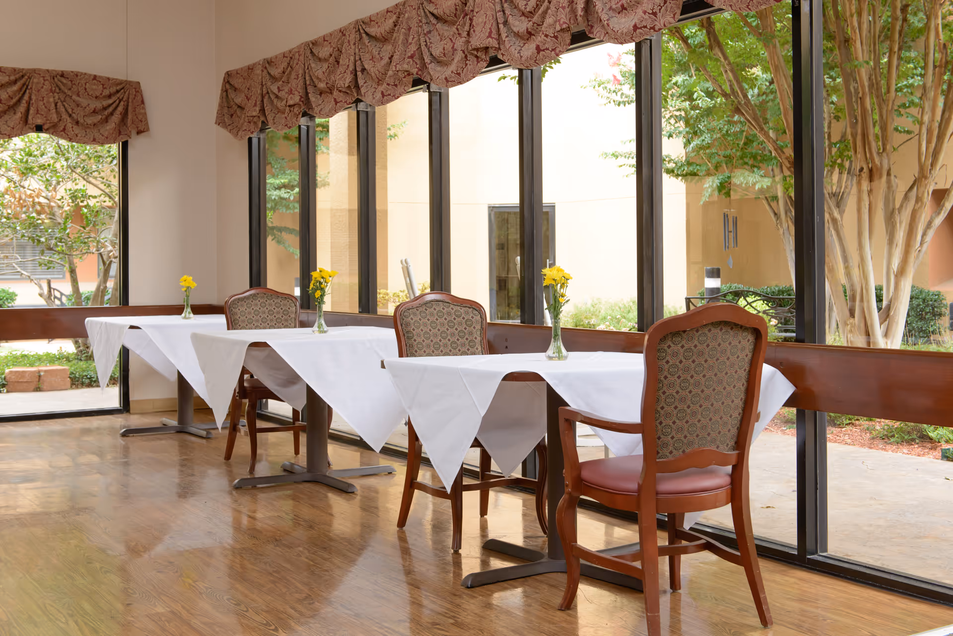A dining area with tables covered in white tablecloths, each adorned with a small vase of yellow flowers. The room has large floor-to-ceiling windows with a view of trees and outdoor greenery. The chairs have wooden frames and patterned upholstery, and the floor is wooden.