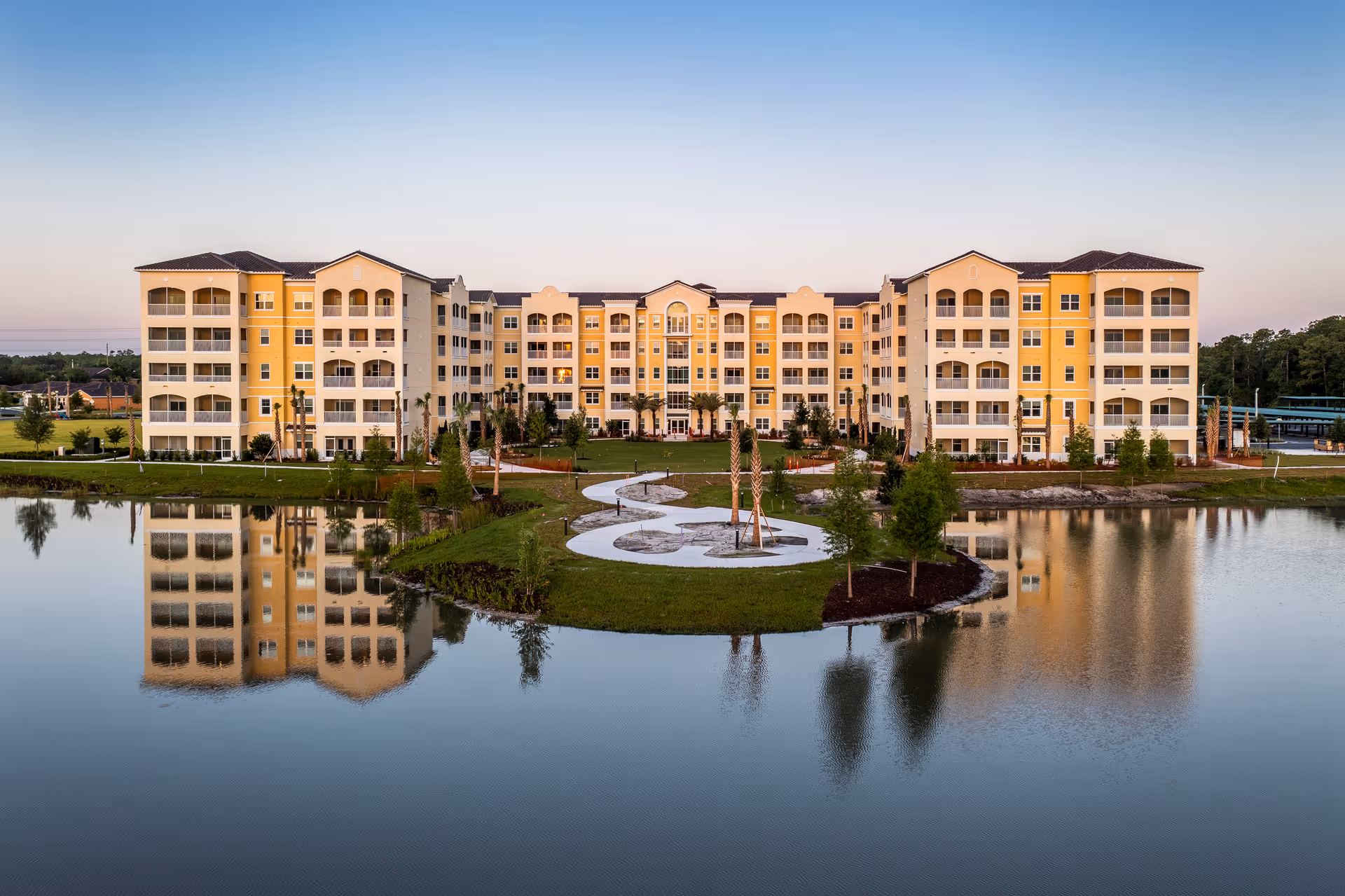 A large, multi-story senior living facility building with yellow and beige exterior walls, multiple balconies, and a tiled roof. The building is reflected in a calm body of water in the foreground, with a landscaped area featuring a winding pathway and young trees between the water and the building. The sky is clear with a gradient from light to deeper blue.