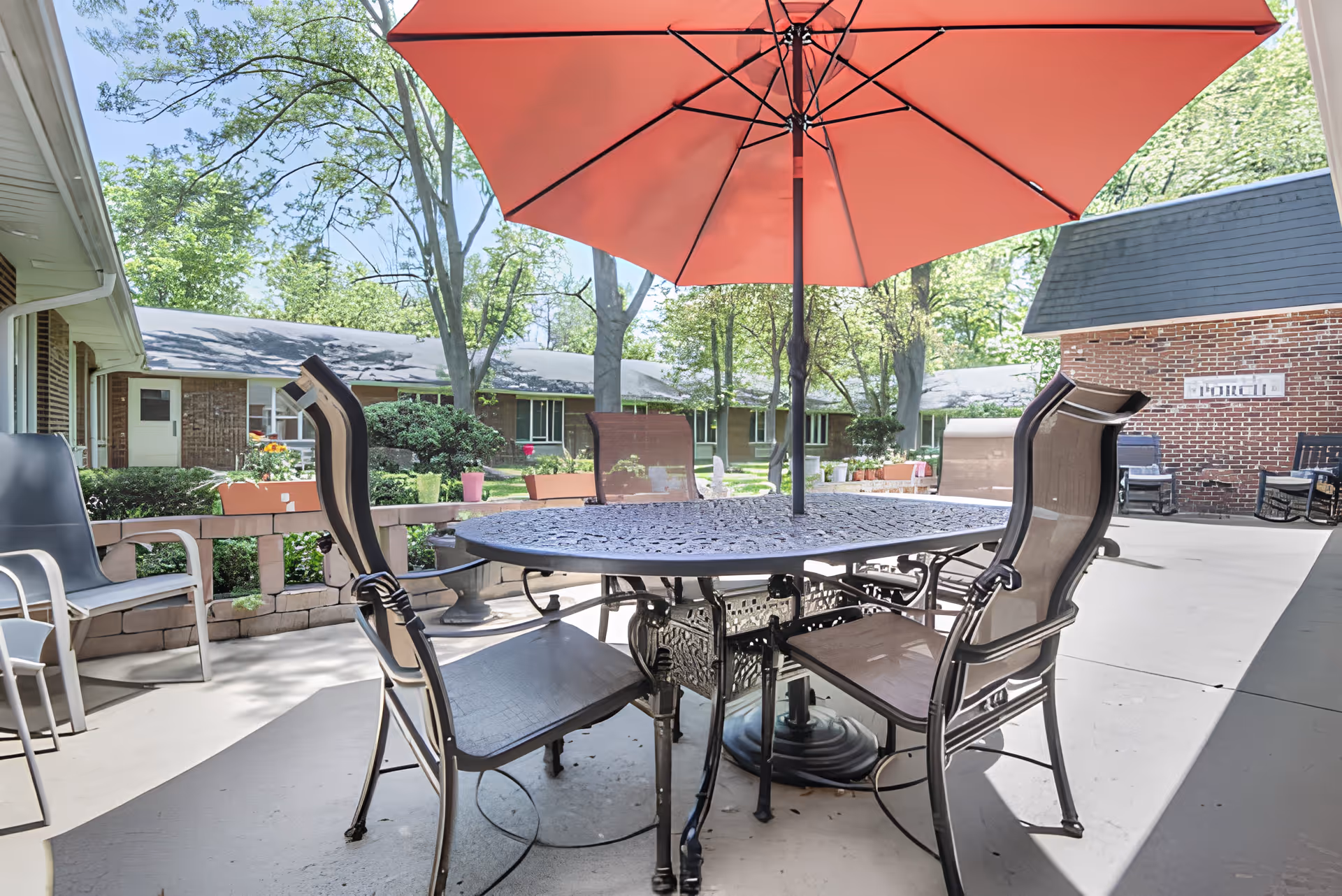 Outdoor patio area with a round metal table and four chairs under a large red umbrella. The patio is surrounded by greenery, trees, and a brick building with a sign that says 'PORCH'.