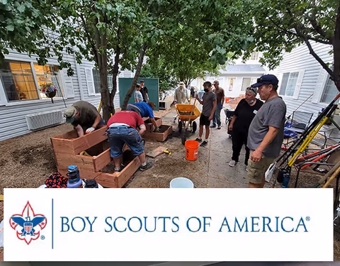 A group of people assembling wooden raised garden beds in a tree-lined courtyard with a Boy Scouts of America banner at the bottom.