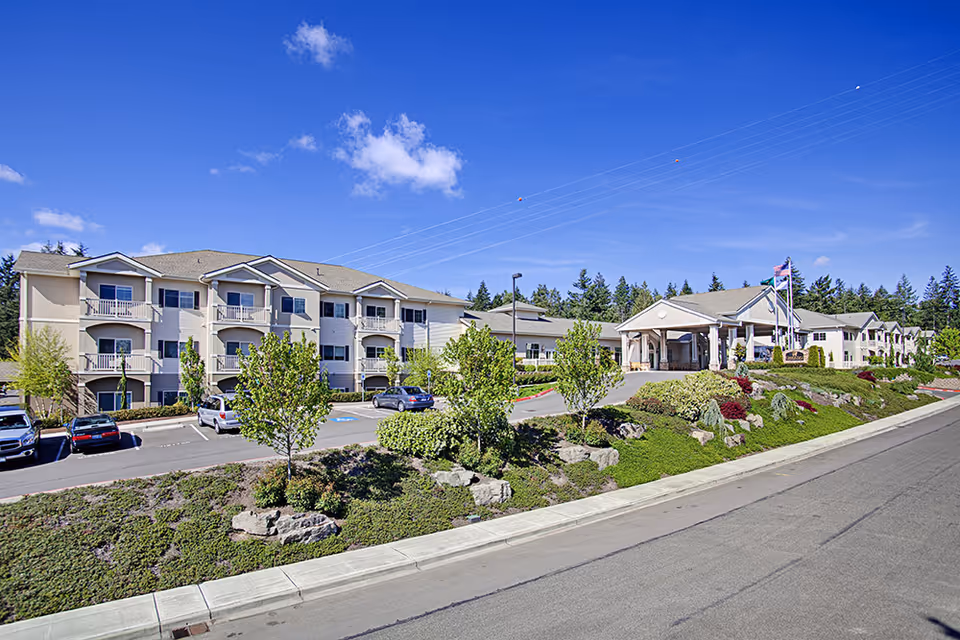 Front exterior of a multi-story senior living facility with balconies, landscaped grounds, parking, and a covered entrance under a blue sky.