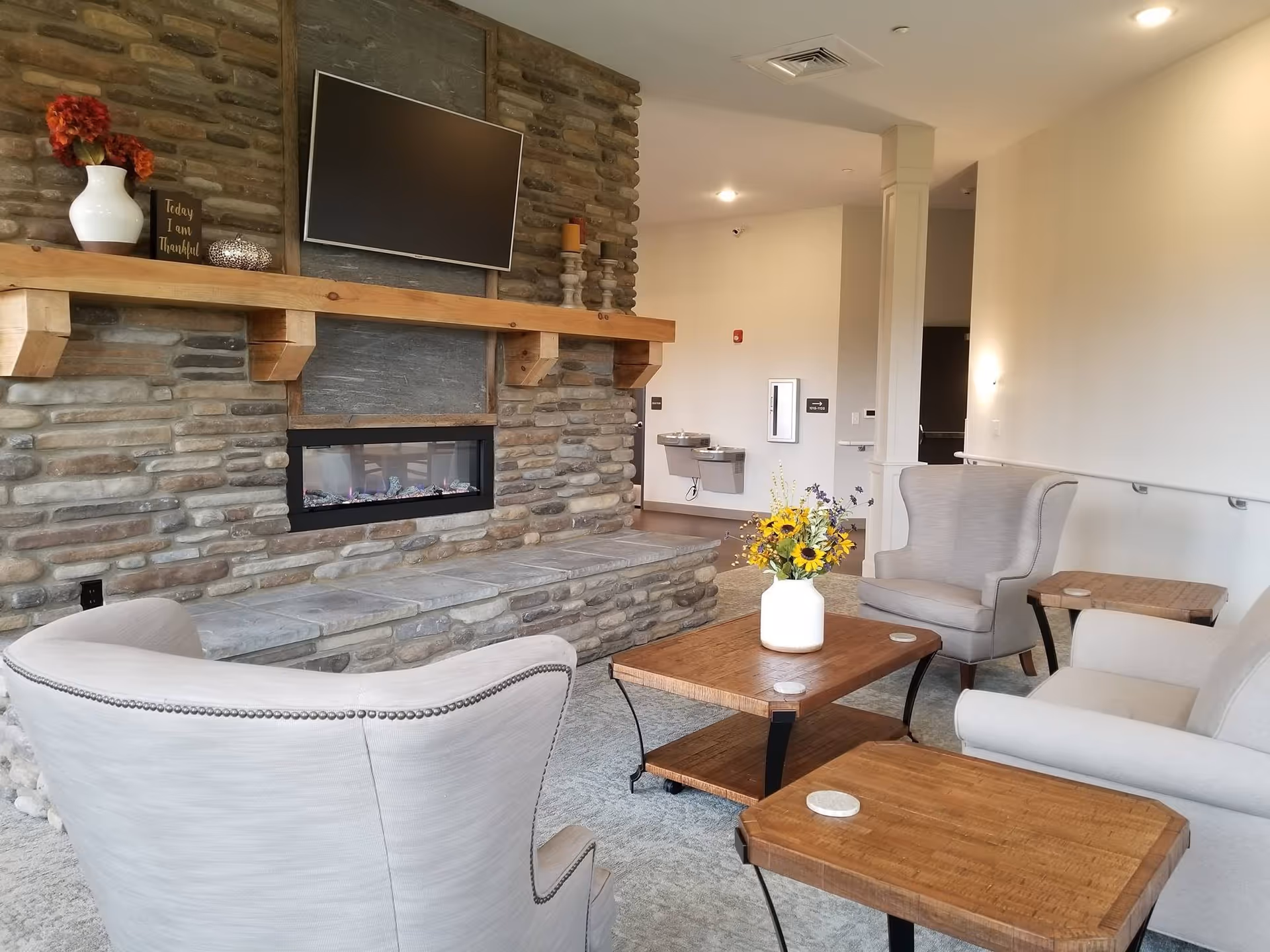 A cozy living room area in an assisted living facility featuring a stone fireplace with a mounted flat-screen TV above it. The room has comfortable beige armchairs and a sofa arranged around wooden coffee and side tables. A vase with yellow and purple flowers sits on the coffee table. The walls are light-colored, and there are water fountains and a fire alarm visible in the background.
