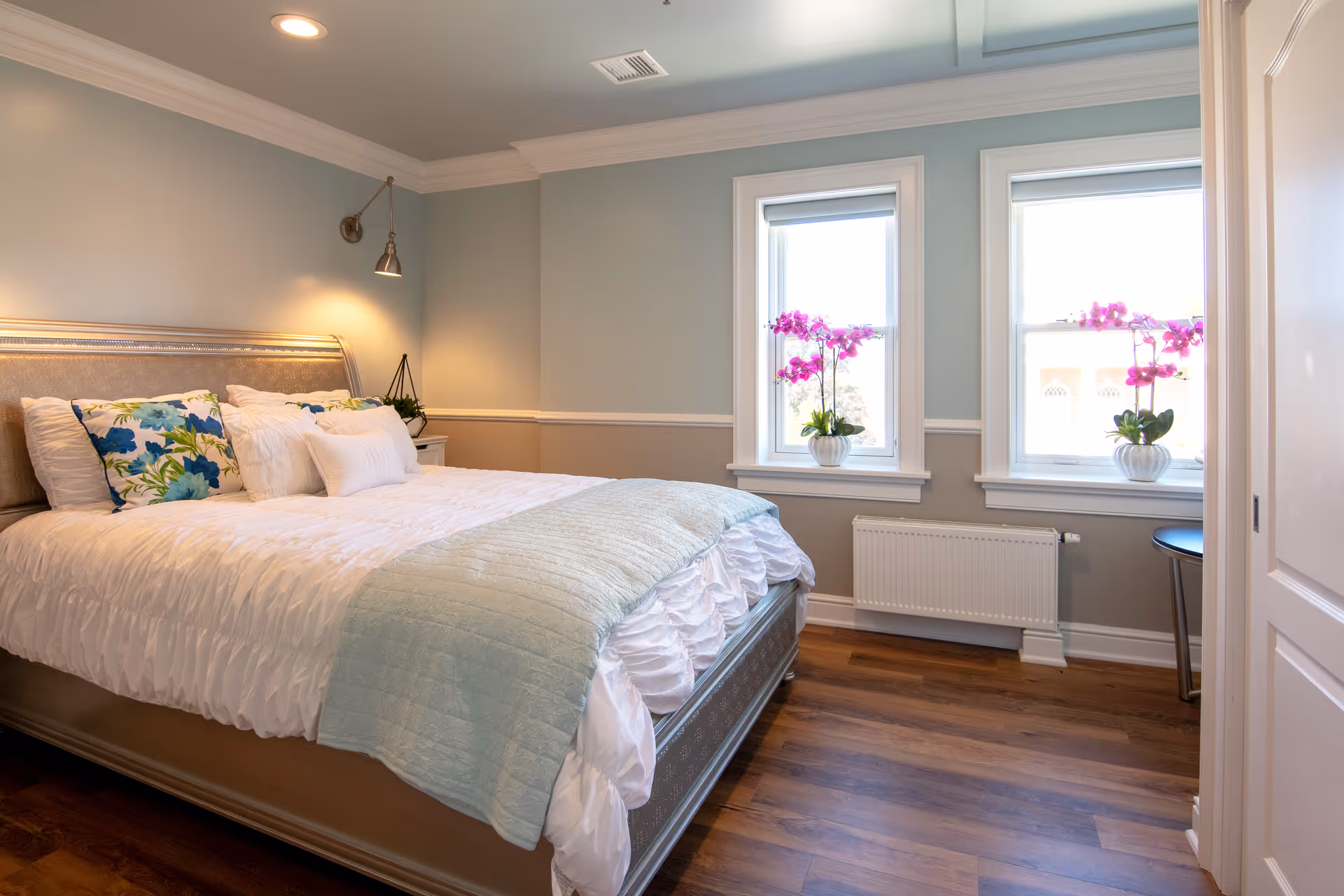 A bright and cozy bedroom with a large bed featuring white and floral pillows, a light blue throw blanket, two windows with pink orchid plants on the windowsills, wooden flooring, and soft blue and beige walls.