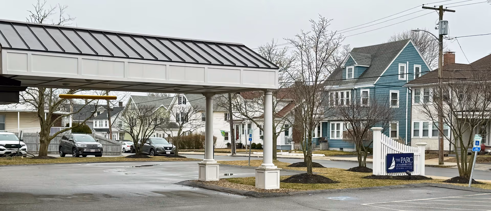 Exterior view of The Parc at Harbor View Senior Living facility showing a covered driveway entrance, parking spaces, leafless trees, and neighboring houses on a cloudy day.