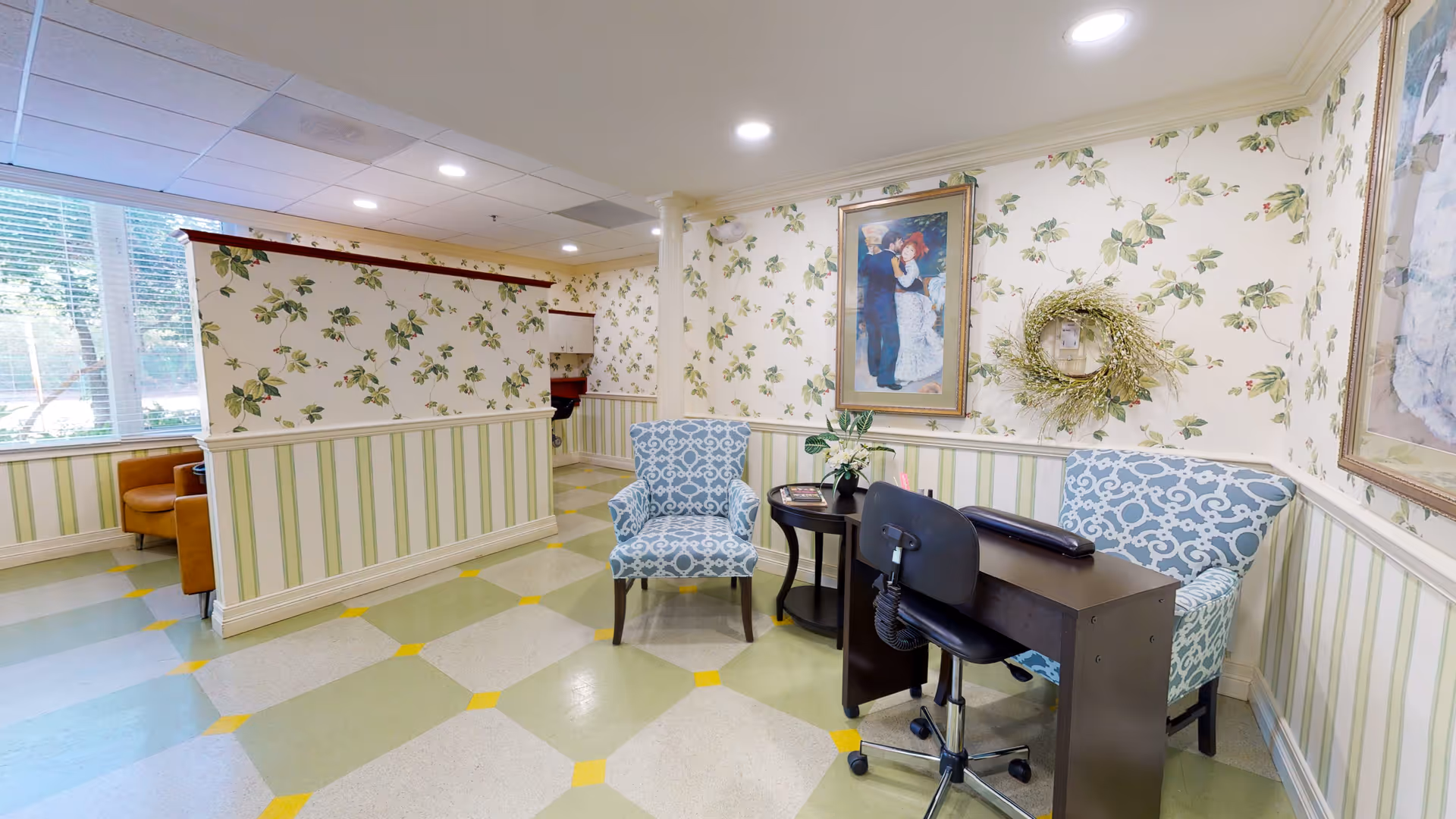 A cozy interior room with floral and striped wallpaper, featuring two blue patterned armchairs, a small dark wooden table with a plant and magazines, and a black office chair behind a dark wooden desk. The floor has a geometric pattern with green, beige, and yellow tiles. There are framed paintings and a decorative wreath on the walls, and a window with blinds letting in natural light.