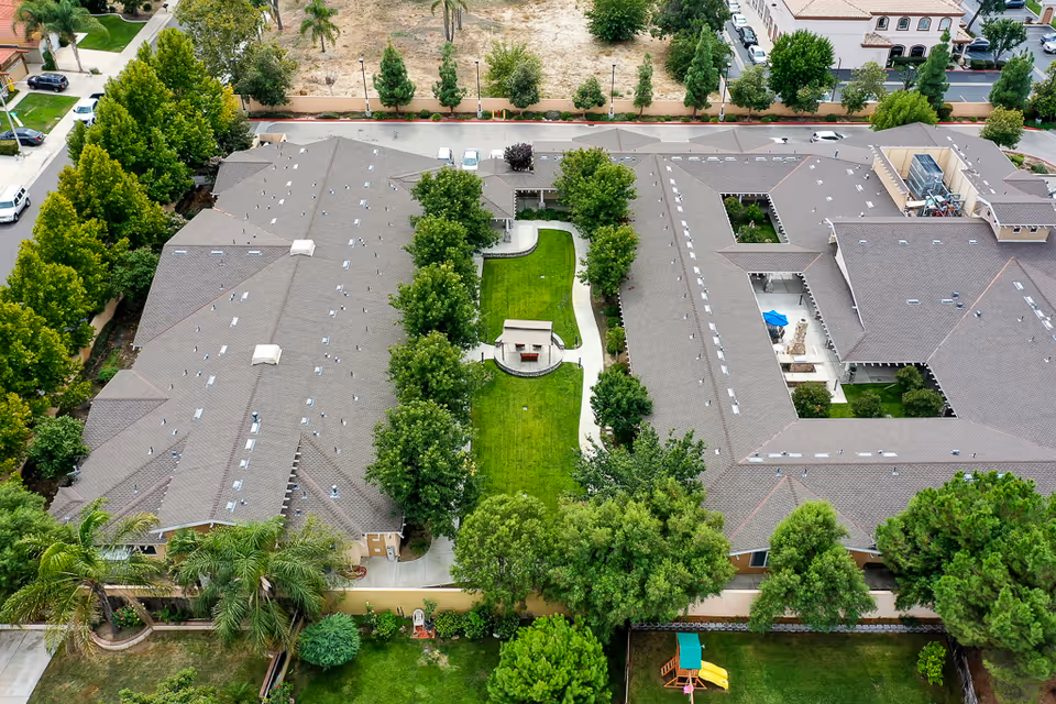 Aerial view of Sunny Rose Senior Living facility showing two large buildings with gray roofs surrounding a green courtyard with trees, a small covered seating area, and pathways. The surrounding area includes trees, a playground with a slide, and nearby streets with parked cars.