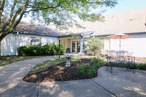 Outdoor courtyard area of a senior living facility with a concrete pathway, garden beds, a birdbath, metal patio table with chairs, and an umbrella. The building has white siding and multiple windows, with trees providing shade.