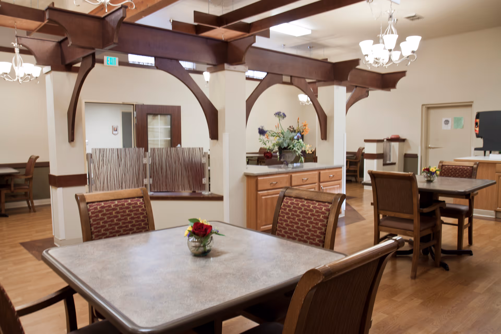Interior view of a dining area in a senior living facility with wooden tables and chairs, a small flower arrangement on each table, decorative wooden beams, and a cabinet with a flower vase. The room has a warm, inviting atmosphere with soft lighting and light-colored walls.
