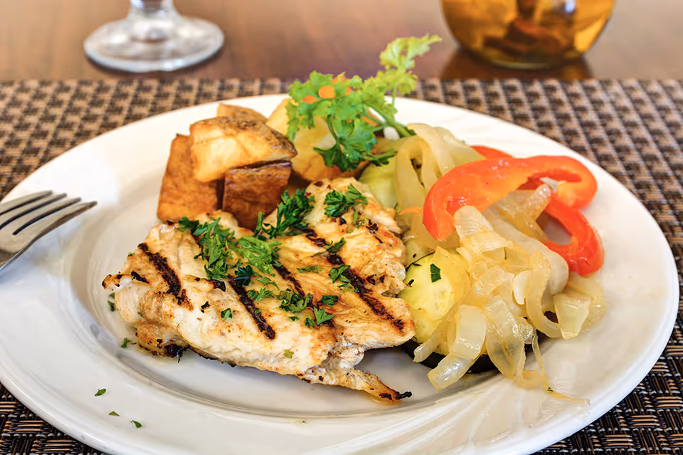 A white plate with grilled chicken garnished with parsley, roasted potatoes, and sautéed onions and peppers on a placemat.