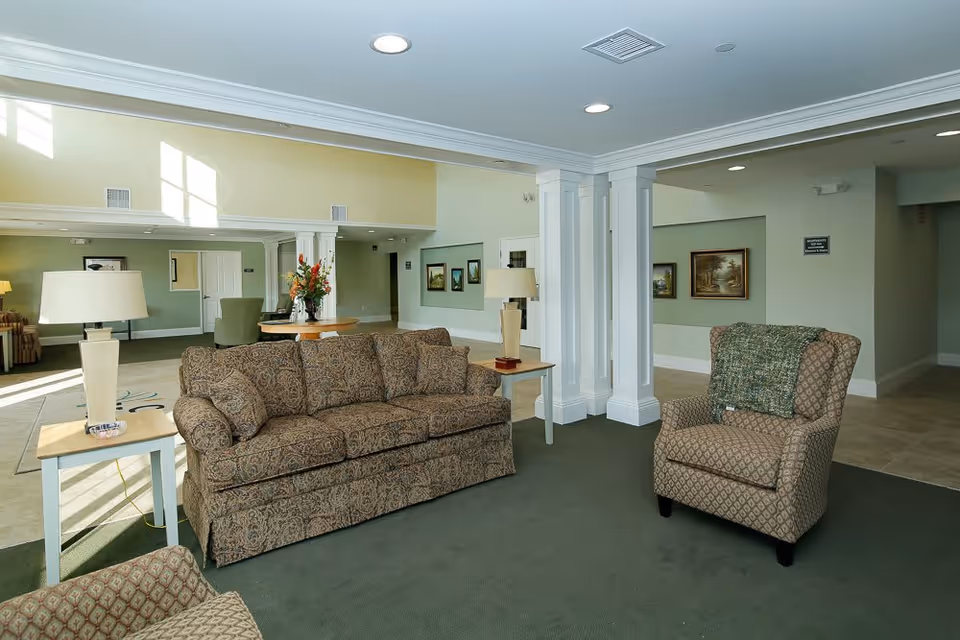 A spacious and well-lit common area with patterned sofas and armchairs arranged on a green carpet. There are two side tables with lamps, a round table with a flower arrangement, and framed paintings on the light green walls. The ceiling has recessed lighting and white columns divide the space.