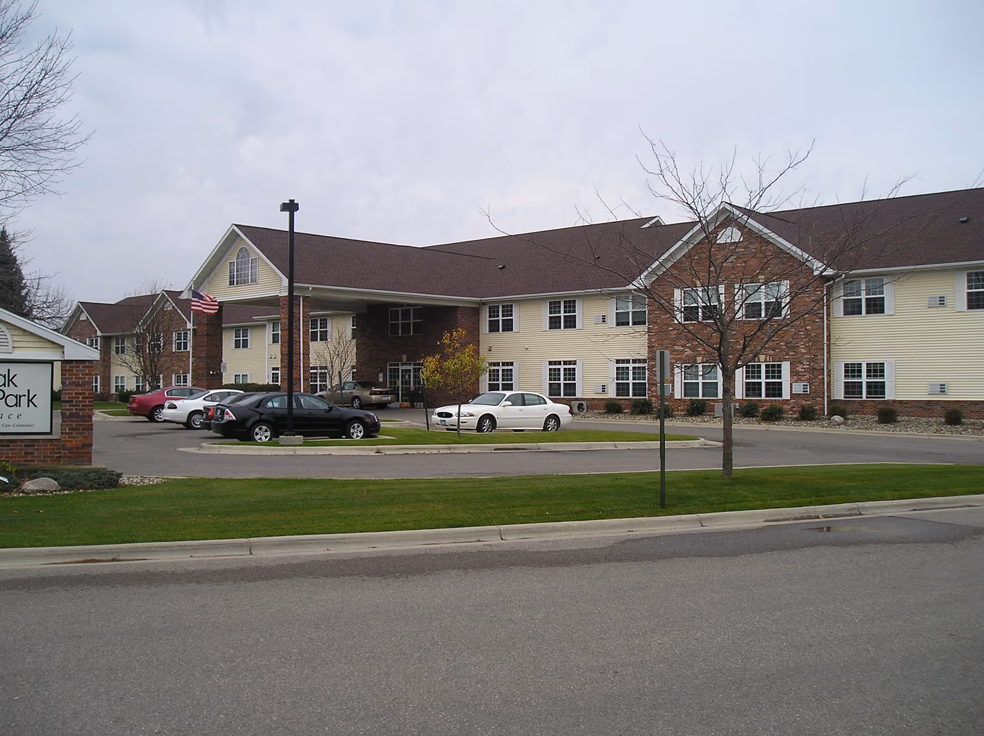 Exterior view of Oak Park Place Albert Lea senior living facility showing a two-story building with beige siding and brick accents, several parked cars, a flagpole with an American flag, and a sign partially visible on the left side.