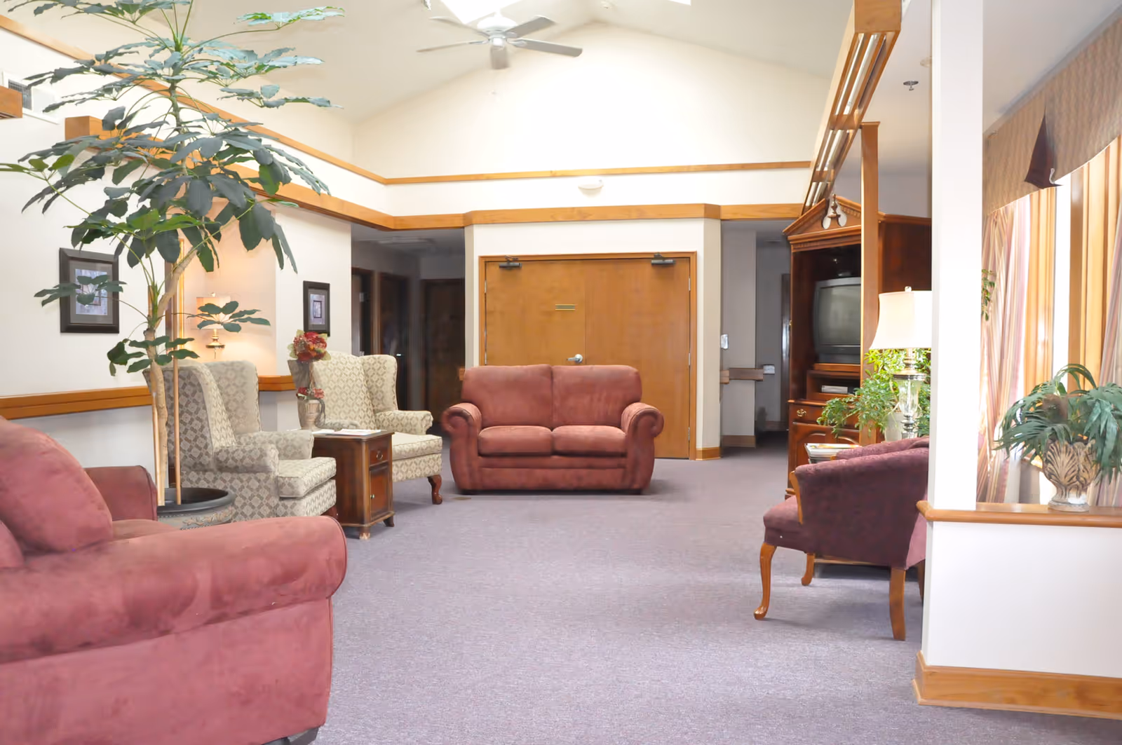 Spacious senior living lounge with upholstered sofas and armchairs, potted plants, and wood-trimmed walls and doors.