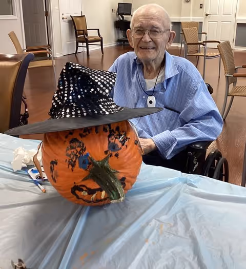 An elderly man in a wheelchair smiling behind a decorated pumpkin wearing a black witch hat with white polka dots, placed on a table covered with a light blue plastic tablecloth in a room with wooden floors and chairs.