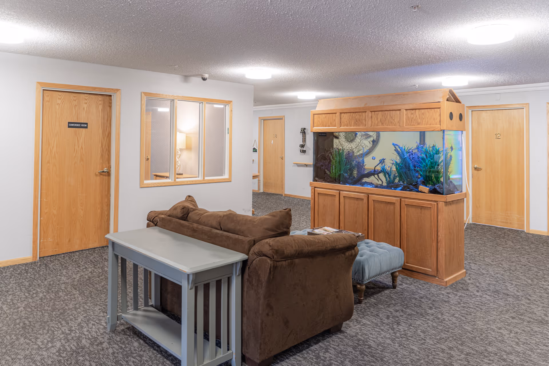 Interior common area of Southern Oaks Place featuring a brown couch with a gray table behind it, a large wooden-framed aquarium filled with plants and fish, a blue cushioned ottoman, and several wooden doors labeled Conference Room, 13, and 12. The walls are light gray with a window showing a lamp inside another room, and the floor is carpeted in a dark pattern.