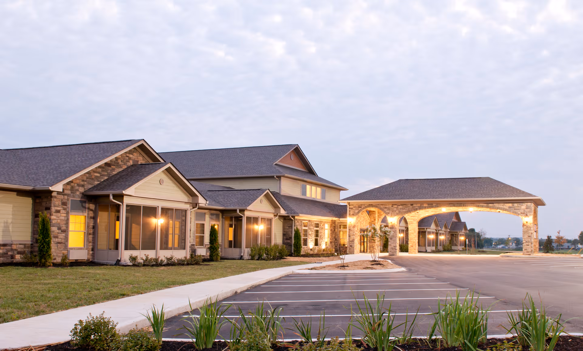 Front exterior of a senior living building with a covered porte-cochere, lit windows, and an empty parking lot.