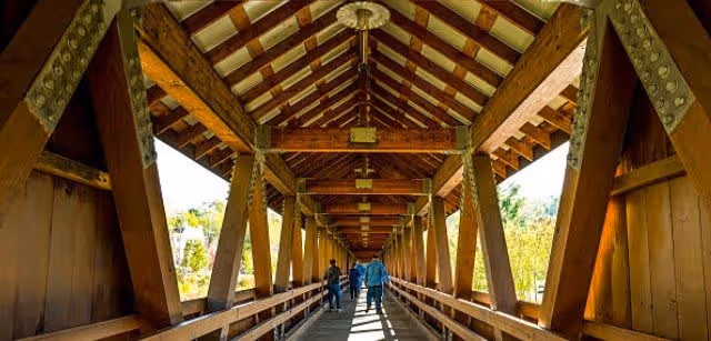A covered wooden bridge with a peaked roof and large wooden beams. Several people are walking along the bridge, and greenery is visible outside on both sides.