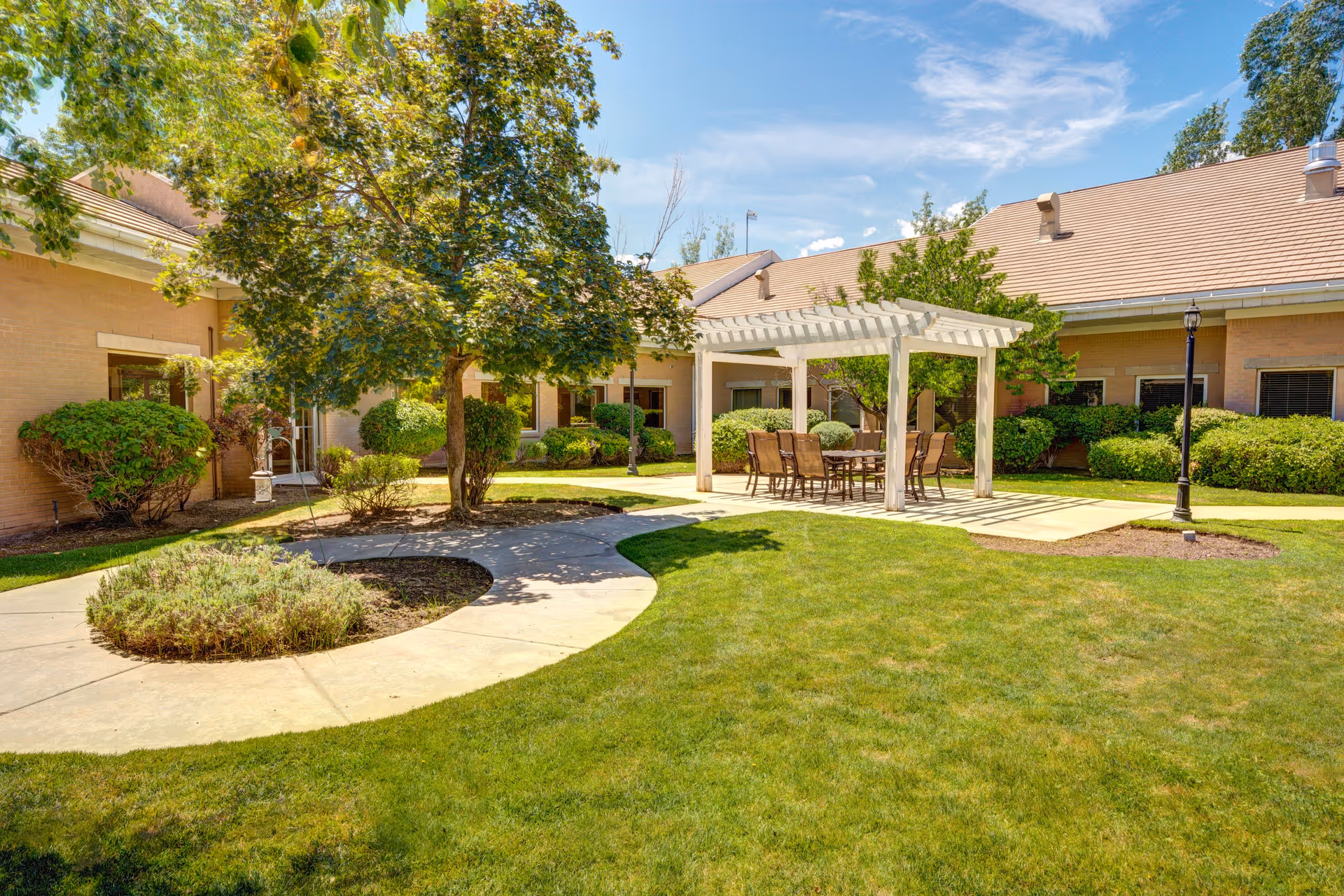Outdoor courtyard area at The Auberge at Aspen Park featuring a green lawn, a large tree, a curved concrete walkway, and a white pergola with a table and several chairs underneath. The building surrounds the courtyard with windows and bushes along the walls.