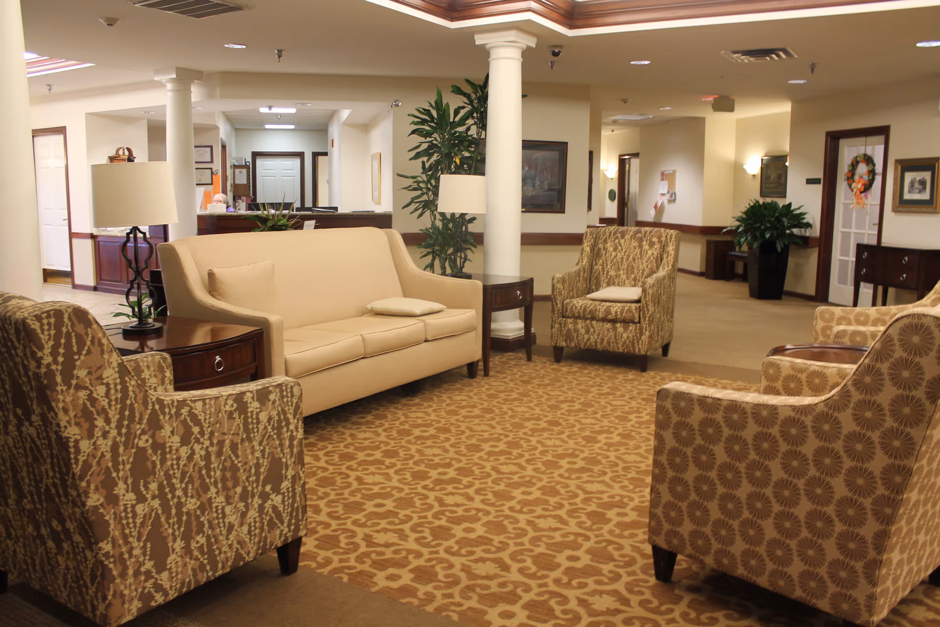 A cozy seating area in a senior living facility with a beige sofa, three patterned armchairs, two side tables with lamps, a patterned carpet, and potted plants. The background shows a reception desk and hallway with doors and framed artwork on the walls.