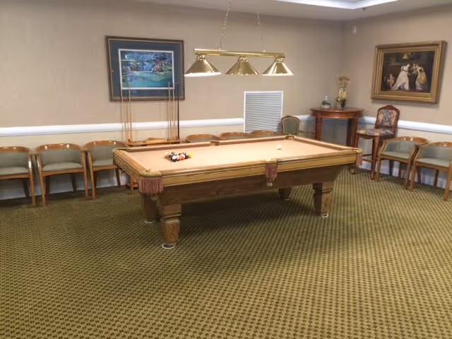 A room with a pool table in the center, surrounded by wooden chairs with green cushions along the walls. The room has patterned green carpet, framed artwork on the walls, and a hanging light fixture with three brass lamps above the pool table.