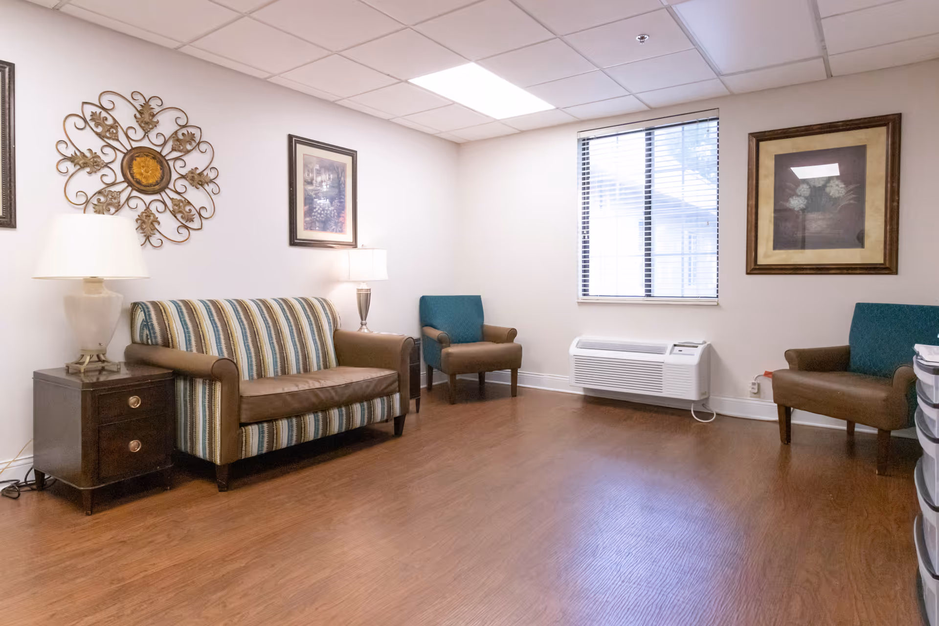 A small sitting area with a striped loveseat and two teal armchairs arranged around the room. There are two table lamps on dark wooden side tables, framed artwork on the walls, a window with blinds, and a wall-mounted air conditioning unit below the window. The floor is wood, and the ceiling has recessed lighting.