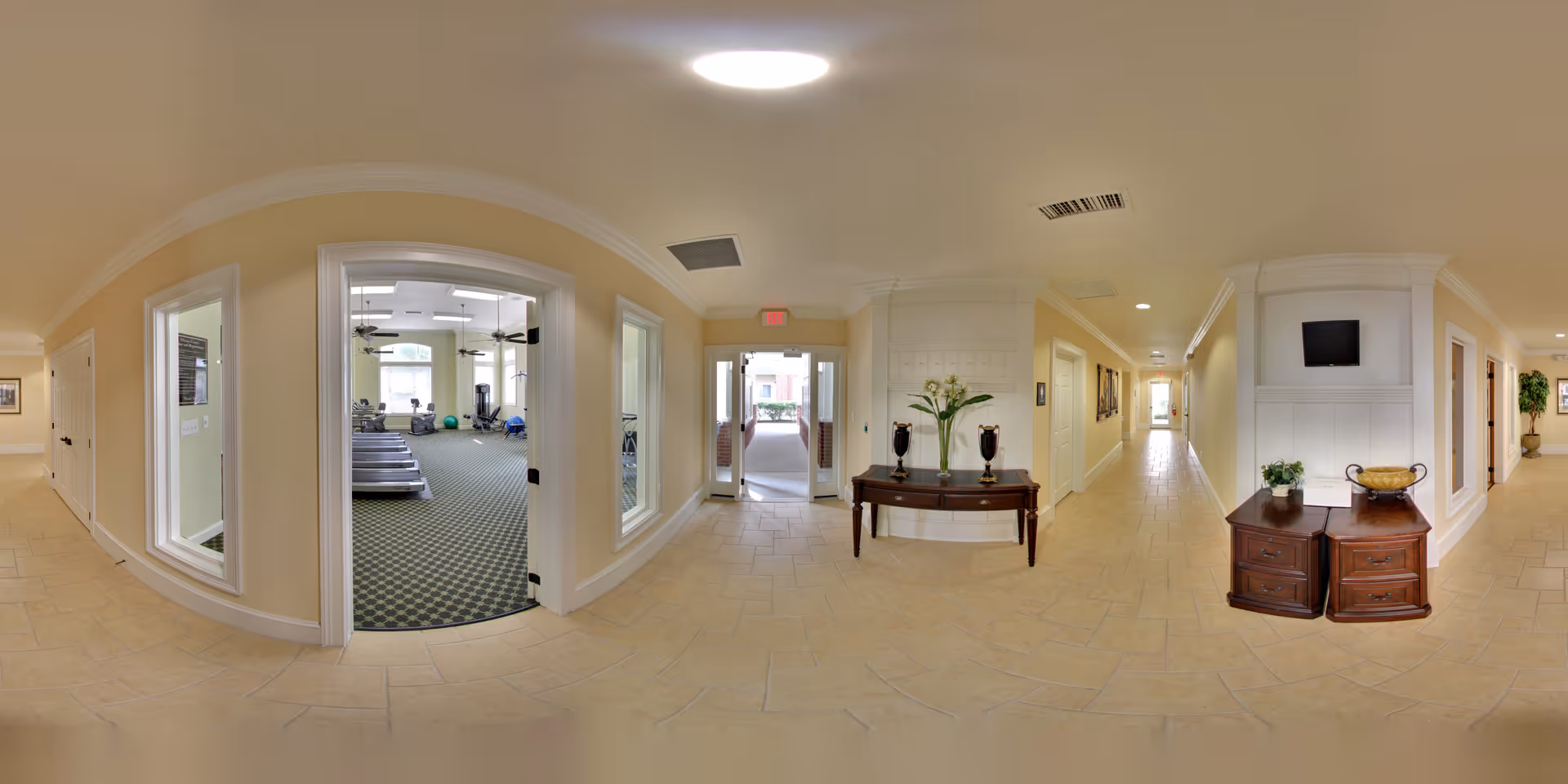 Wide-angle view of a hallway in a senior living facility with beige walls and tiled floors. On the left, there is a room with exercise equipment including treadmills and exercise balls visible through large windows. In the center, a wooden table with decorative vases and flowers is placed against the wall. The hallway extends to the right with additional furniture, including a small cabinet with a plant and a bowl, and a mounted TV above it. The hallway is well-lit with ceiling lights and has multiple doorways leading to other rooms.
