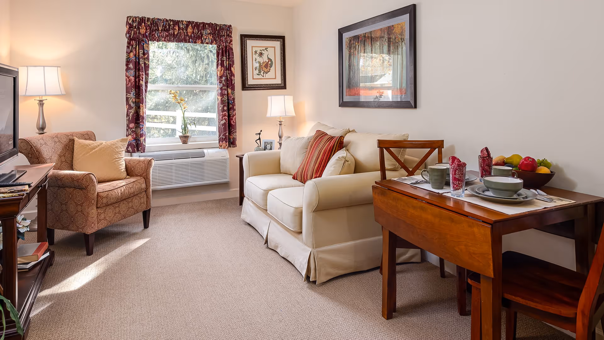 A cozy living room area with a beige loveseat adorned with red and beige pillows, a patterned armchair with a beige pillow, a wooden side table with a lamp, and a wooden dining table set for two with dishes, cups, and a bowl of fruit. A window with floral curtains lets in natural light, and framed artwork hangs on the walls.