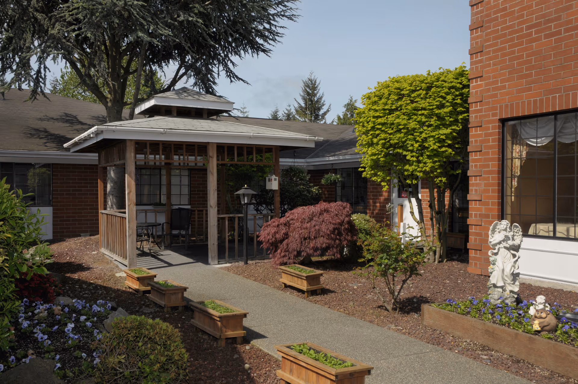 Outdoor garden area at Hallmark Manor featuring a paved walkway lined with wooden planters, a small wooden gazebo with chairs underneath, various shrubs and trees, and a decorative angel statue near a building with large windows.