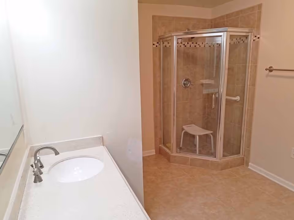 Bathroom with a corner glass shower enclosure containing a shower stool and grab bar, and a white vanity sink with faucet.