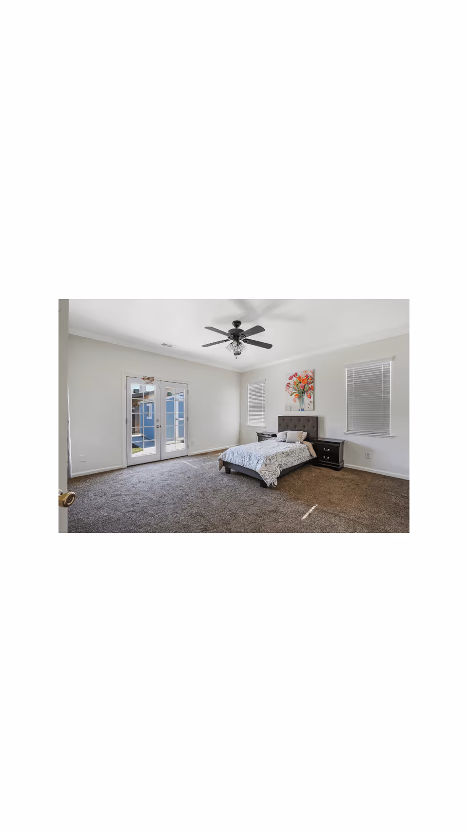 A spacious bedroom with a large bed covered in a white and gray patterned comforter, two black nightstands on either side of the bed, a ceiling fan with lights, two windows with blinds, a colorful floral painting above the bed, and double glass doors leading outside.