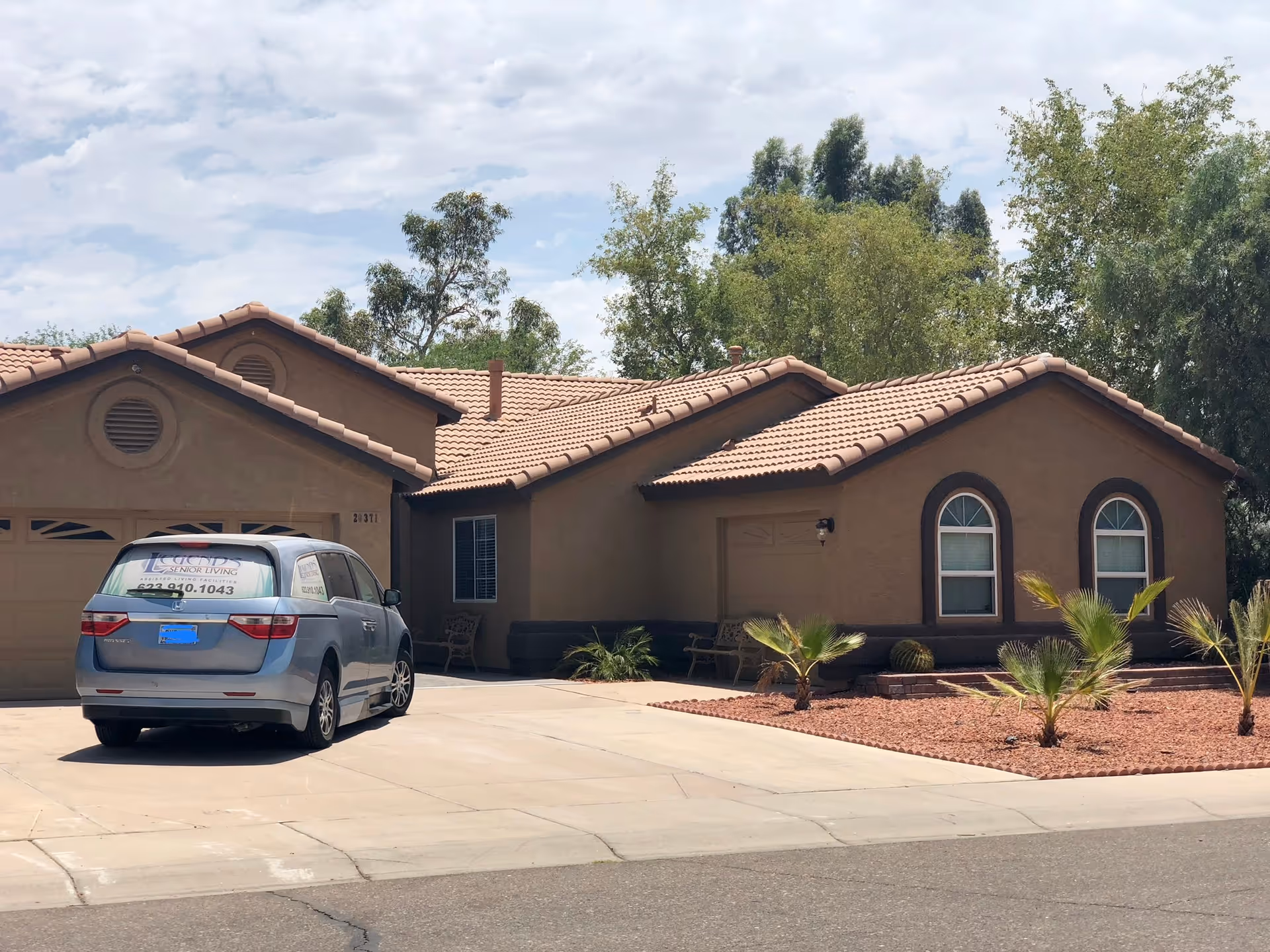 Front exterior of a single-story tan stucco house with a tiled roof, driveway, desert landscaping, and a van parked in the driveway.