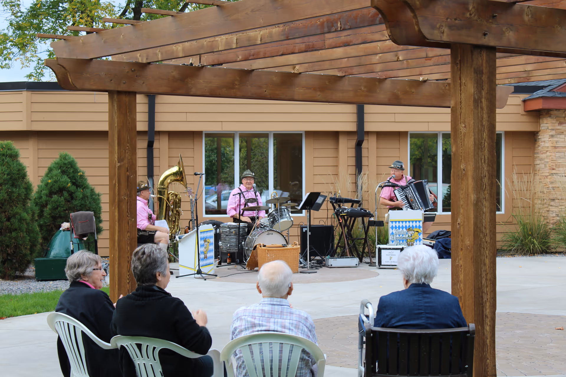 Seniors seated under a wooden pergola watching a small band perform in an outdoor courtyard.