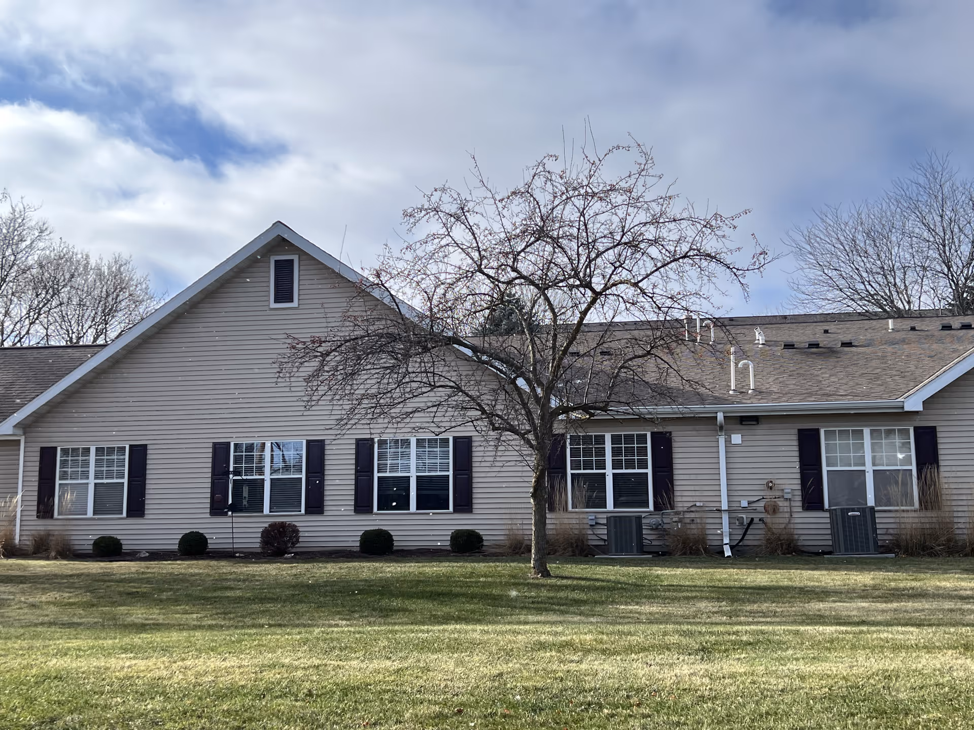 Exterior view of a single-story residential building with beige siding, several windows with dark shutters, a leafless tree in front, and a well-maintained grassy lawn under a partly cloudy sky.