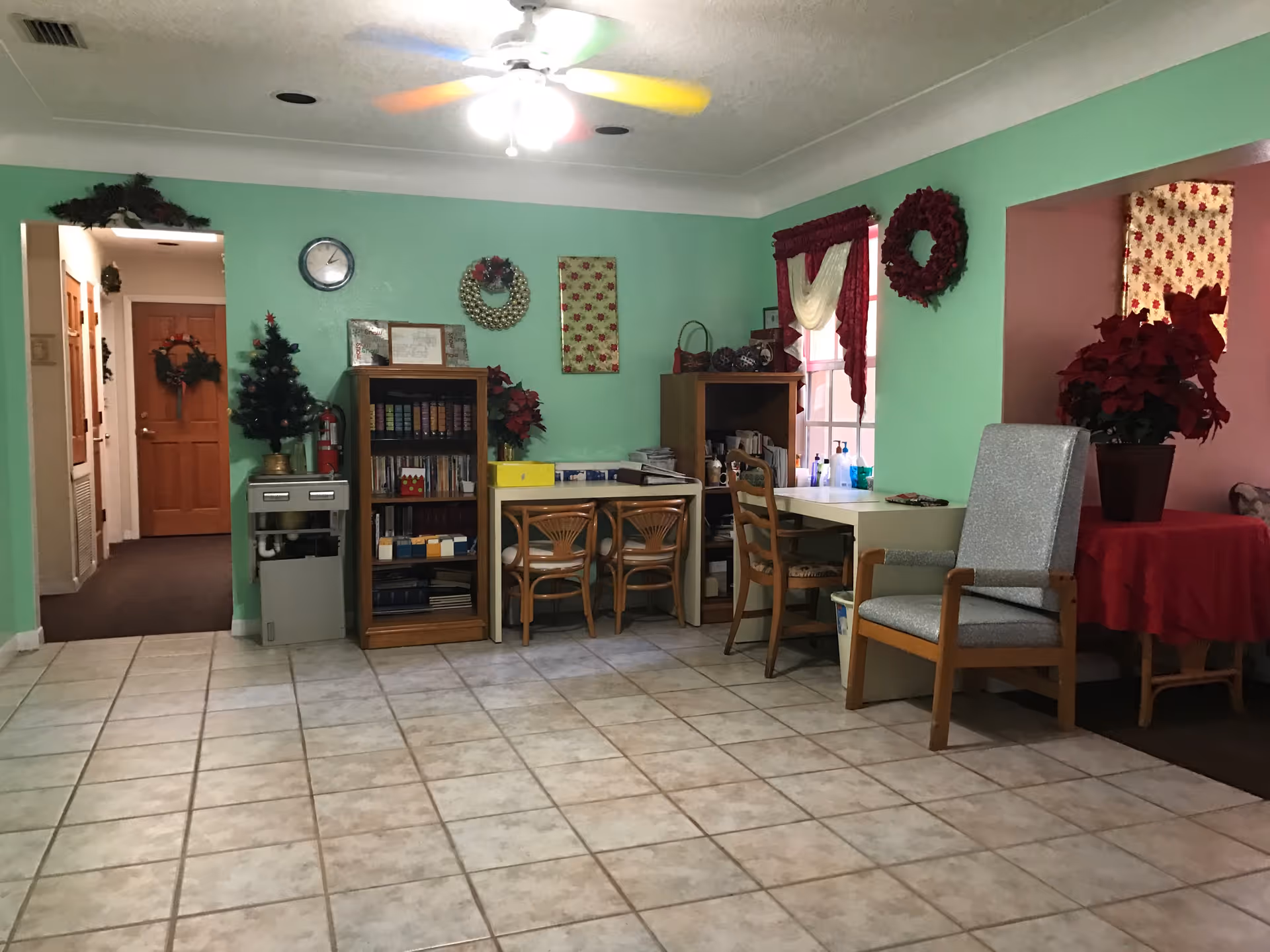 Interior room with light green walls and tiled floor, featuring a small decorated Christmas tree on a cabinet, bookshelves, a clock on the wall, a table with chairs, a comfortable armchair, and holiday wreaths and poinsettia plants as decorations.