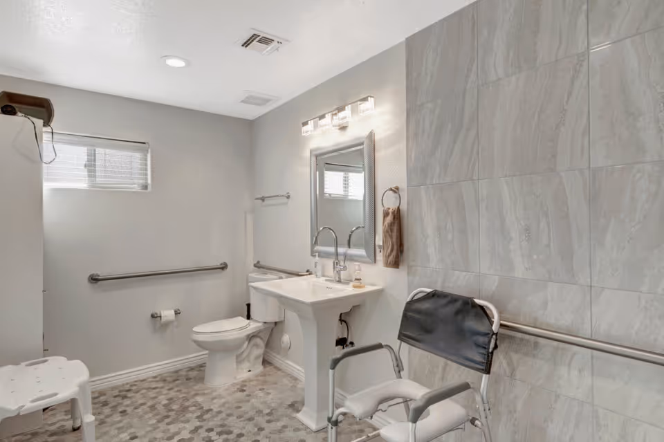 A clean, accessible bathroom with a white toilet, pedestal sink, and a large mirror above the sink. The bathroom features grab bars on the walls, a shower chair, and a tiled wall with a gray pattern. There is a small window with blinds letting in natural light.