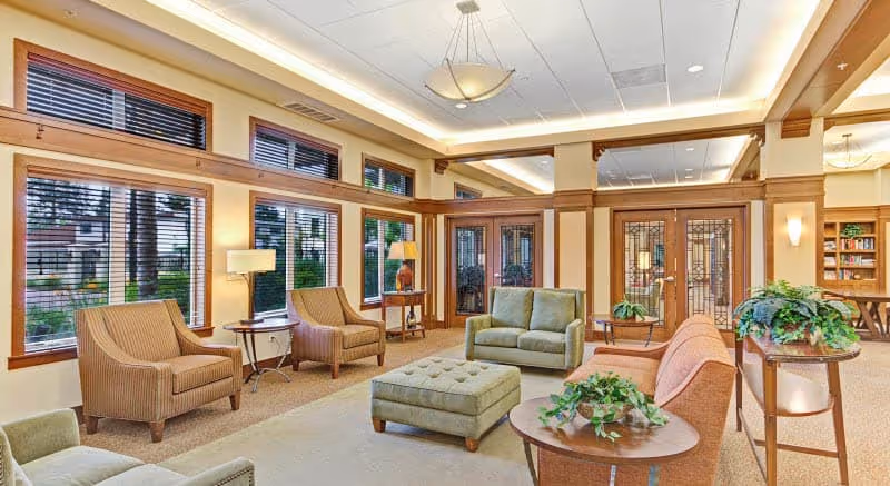 A bright and spacious living room area in a senior living facility with large windows, several upholstered chairs and sofas arranged around a central ottoman, wooden side tables with lamps, and decorative plants. The room features warm wood trim and glass-paneled doors leading to another area.