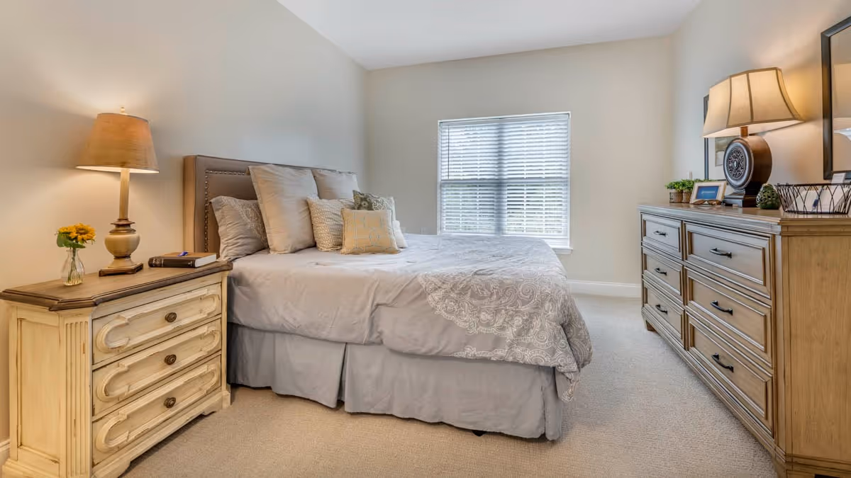 A cozy bedroom with a neatly made bed featuring multiple pillows and a patterned comforter. To the left of the bed is a wooden nightstand with a lamp, a book, and a small vase with a sunflower. On the right side of the room is a wooden dresser with a lamp, a clock, framed photos, and decorative items. A window with blinds is centered on the far wall, allowing natural light to brighten the room.