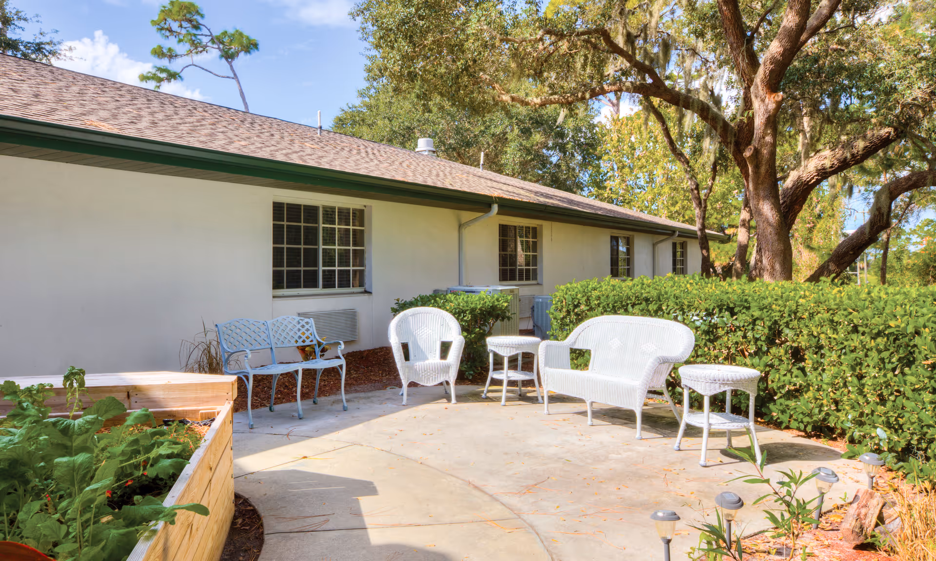 Outdoor patio area at Brookdale Tarpon Springs with white wicker chairs, a small table, a metal bench, and a raised garden bed with plants. The patio is adjacent to a building with windows and surrounded by green bushes and large trees under a blue sky.
