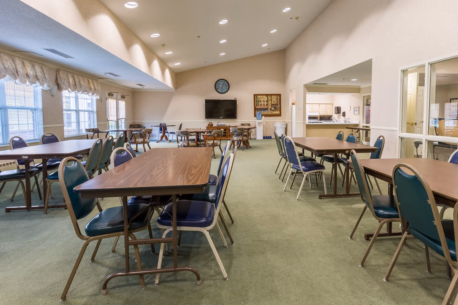 A spacious dining room with multiple rectangular tables and green cushioned chairs arranged around them. Large windows with valances allow natural light to fill the room. A wall-mounted TV and a clock are visible on the far wall, along with a water dispenser and framed artwork. The room has a green carpet and beige walls, with an open doorway leading to a kitchen area.