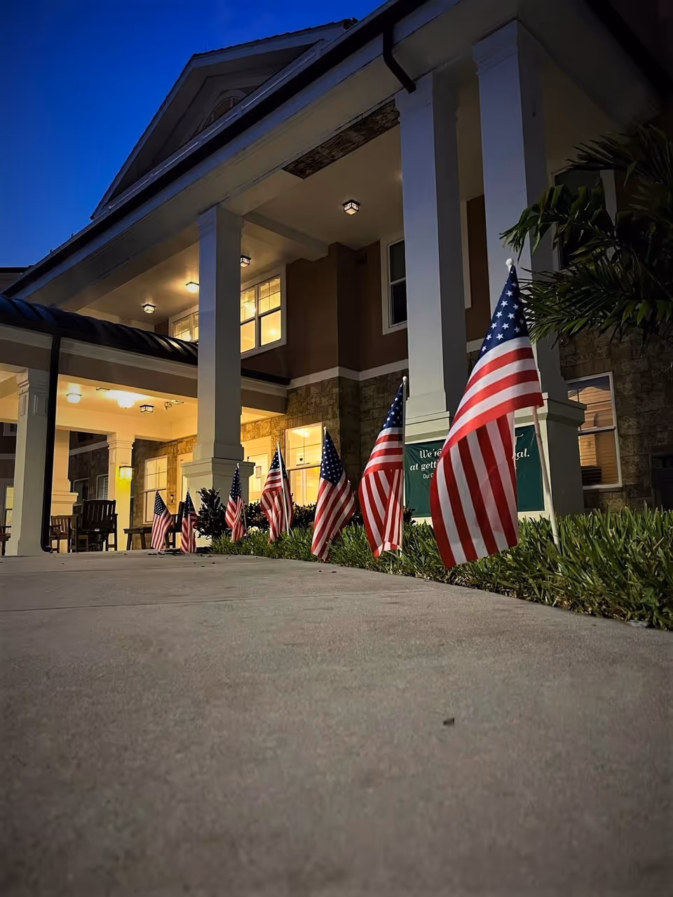 Front entrance of a senior living building at dusk with a row of American flags lining the walkway and illuminated porch columns.