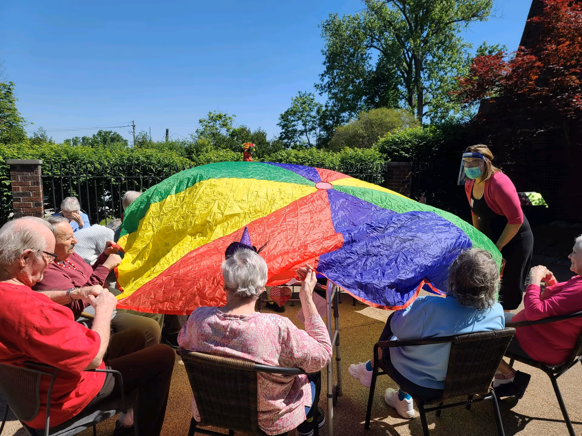 A group of elderly people sitting in chairs outdoors, holding the edges of a large colorful parachute. A caregiver wearing a face shield and mask is standing nearby. The setting is a sunny day with trees and bushes in the background.