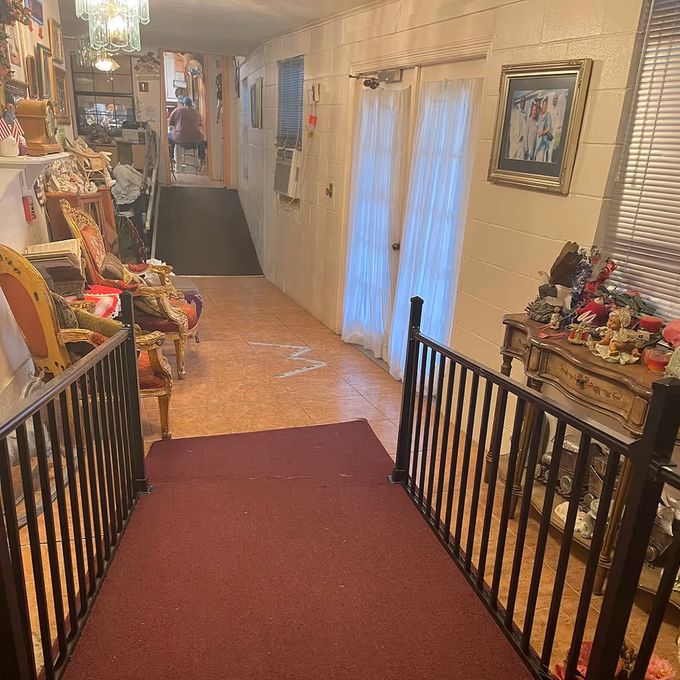 Interior hallway of White's Cottage Center with a maroon carpeted ramp and black railings leading into a corridor with tile flooring. The left side features ornate chairs and various decorative items, while the right side has a wooden table with figurines and a framed picture on the wall. A person is seated at the far end of the hallway.