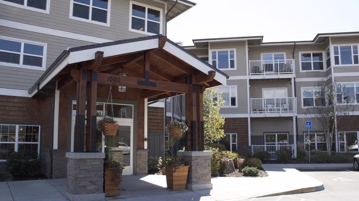 Exterior view of a senior living facility entrance with a covered wooden porch supported by stone pillars. The building has multiple windows and balconies, with landscaping including small trees and shrubs around the entrance area.