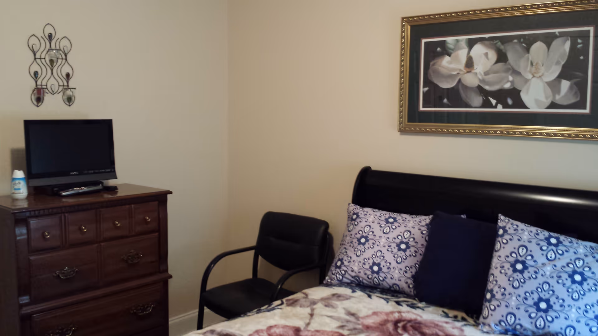 A bedroom with a bed featuring floral-patterned bedding and three pillows, a black chair next to the bed, a wooden dresser with a small TV and remote on top, a decorative wall candle holder, and a framed picture of white flowers on the wall.