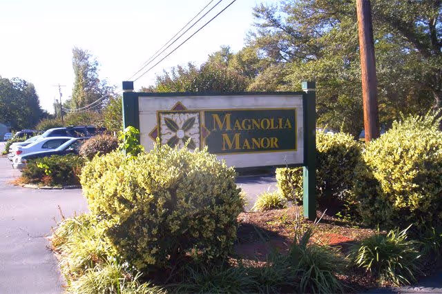 Outdoor view of a Magnolia Manor sign surrounded by bushes and greenery with parked cars and trees in the background under a clear sky.