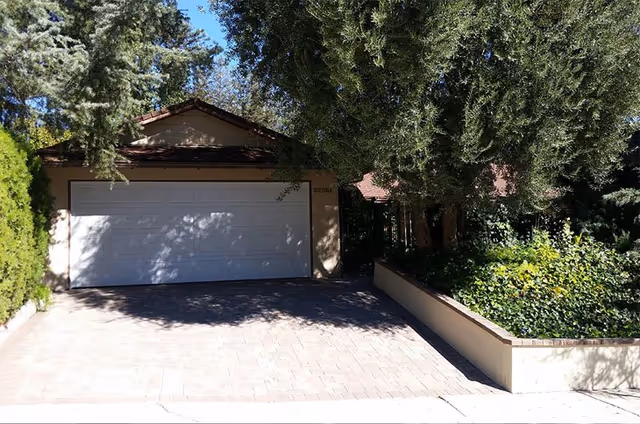 A driveway leading to a closed white garage door attached to a beige house, surrounded by trees and greenery on both sides.