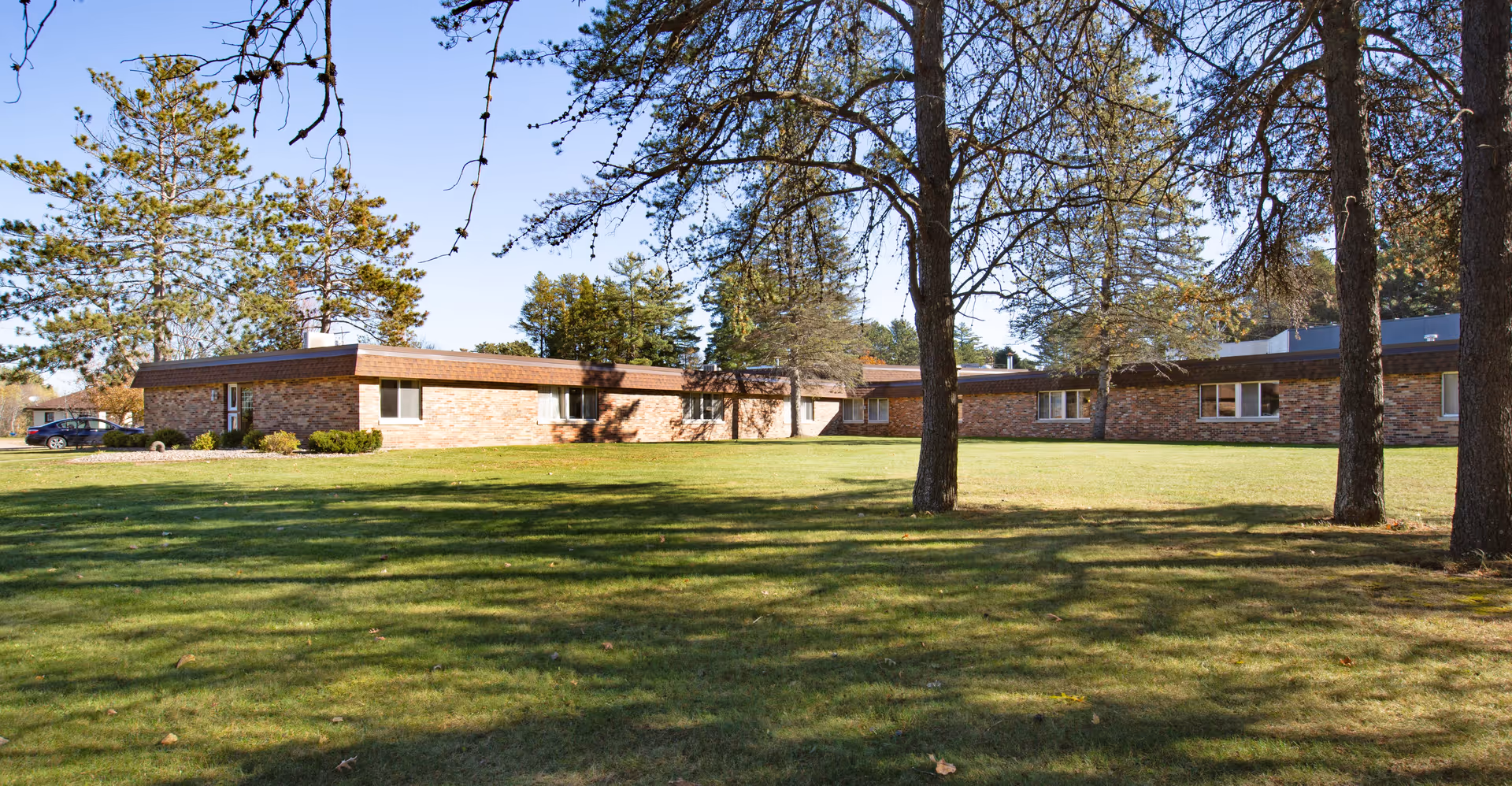 Single-story brick senior living facility building with a large grassy lawn and trees under a clear sky.