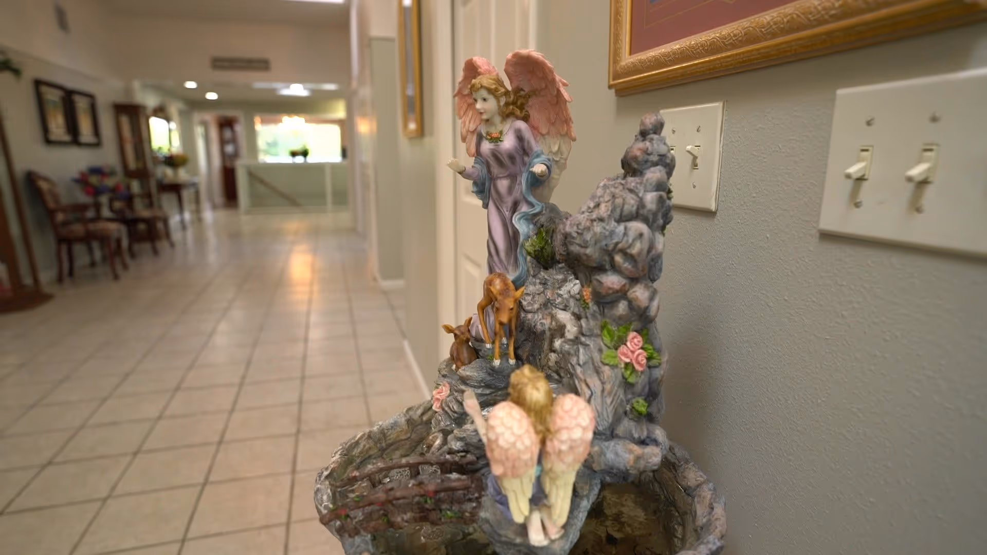 A hallway with tiled floor and light-colored walls, decorated with framed pictures and chairs along the left side. In the foreground on the right side, there is a decorative figurine of two angels with pink wings and two small deer, set on a rock-like base. Light switches are visible on the wall behind the figurine.