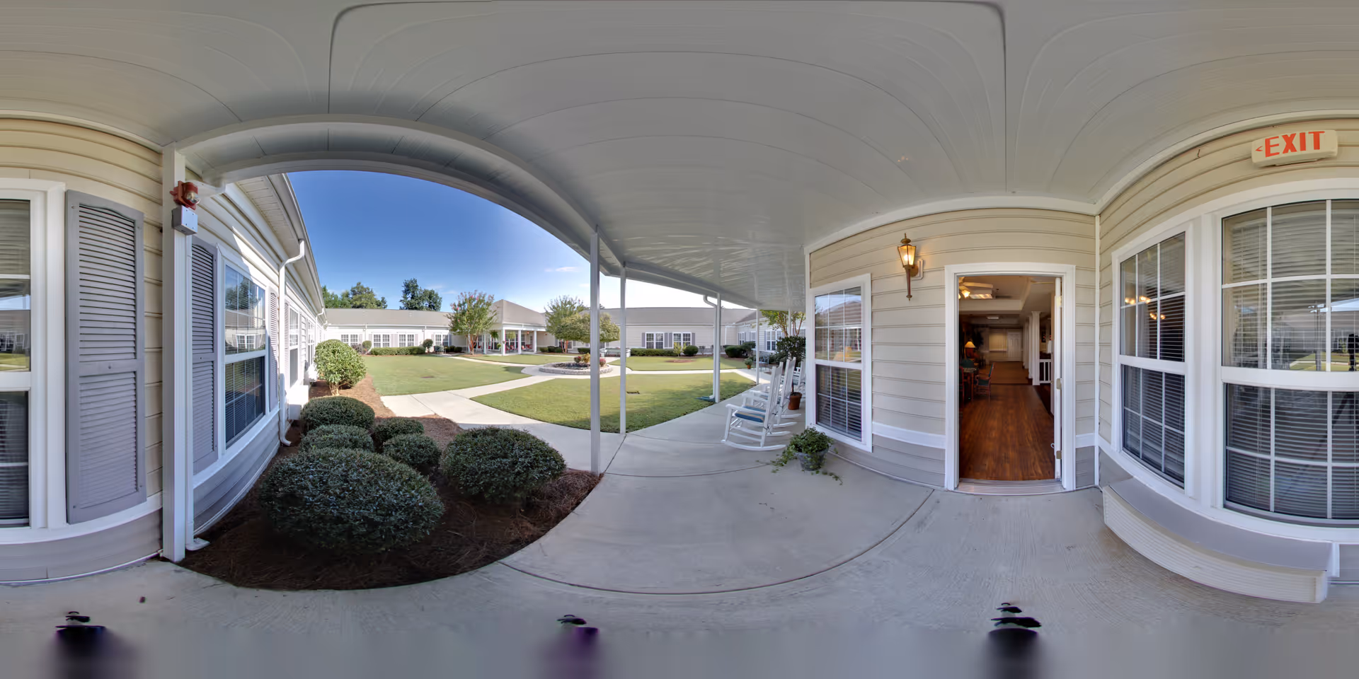 Covered outdoor walkway with white rocking chairs and bushes along the side, leading to a courtyard with green grass and trees surrounded by single-story buildings under a clear blue sky.