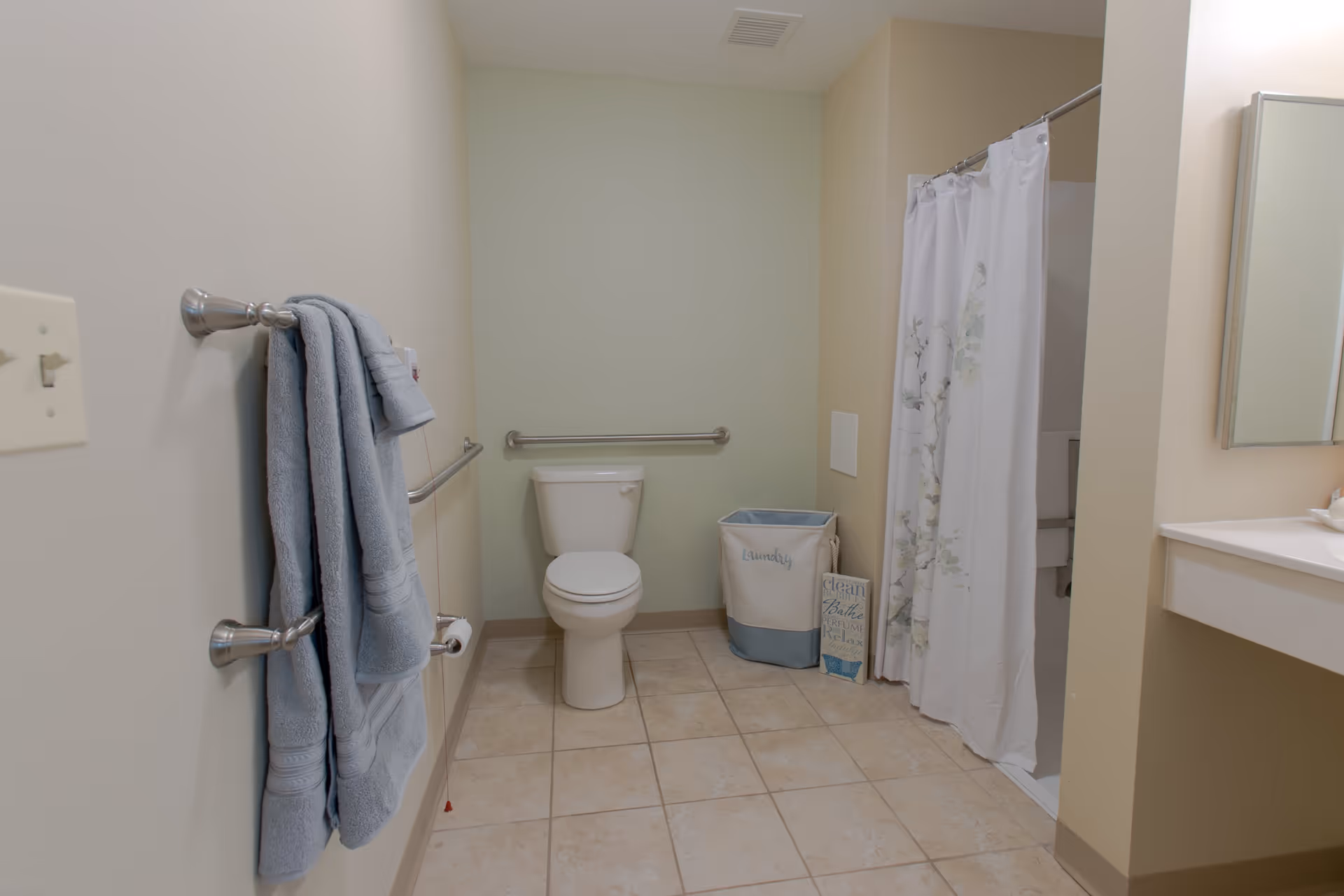 A clean bathroom with beige tiled floor and light-colored walls. There is a white toilet centered against the back wall with a metal grab bar behind it. To the right is a shower with a white curtain featuring a subtle floral design. A laundry basket and a decorative sign are placed next to the shower. On the left wall, there are two towel bars holding folded blue towels. A portion of a white countertop and mirror is visible on the right side of the image.