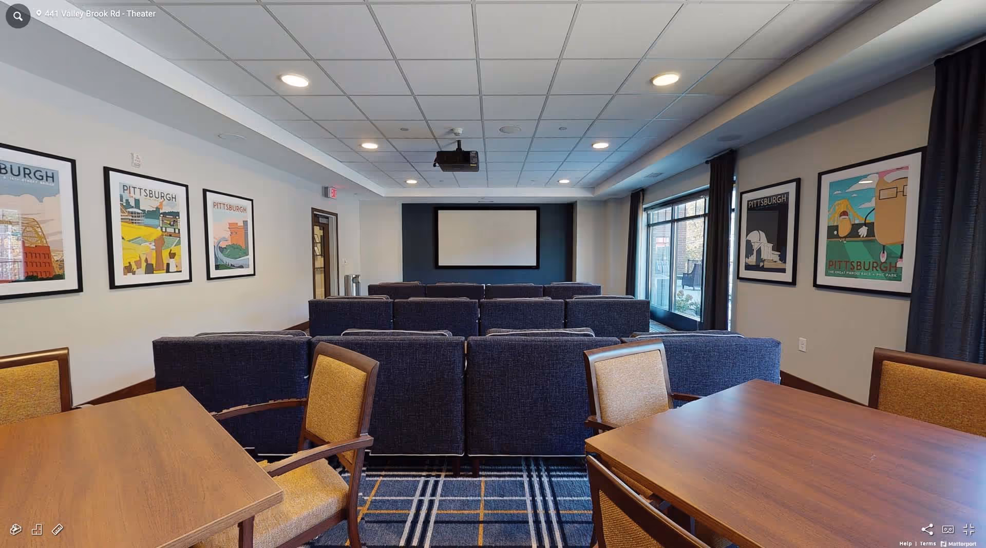 Interior view of a theater room with rows of blue cushioned chairs facing a large blank screen mounted on a dark accent wall. The room has a white ceiling with recessed lighting and a projector hanging from the center. On the left and right walls, there are framed Pittsburgh-themed posters. In the foreground, there are wooden tables with yellow upholstered chairs. Large windows with dark curtains are on the right side of the room.